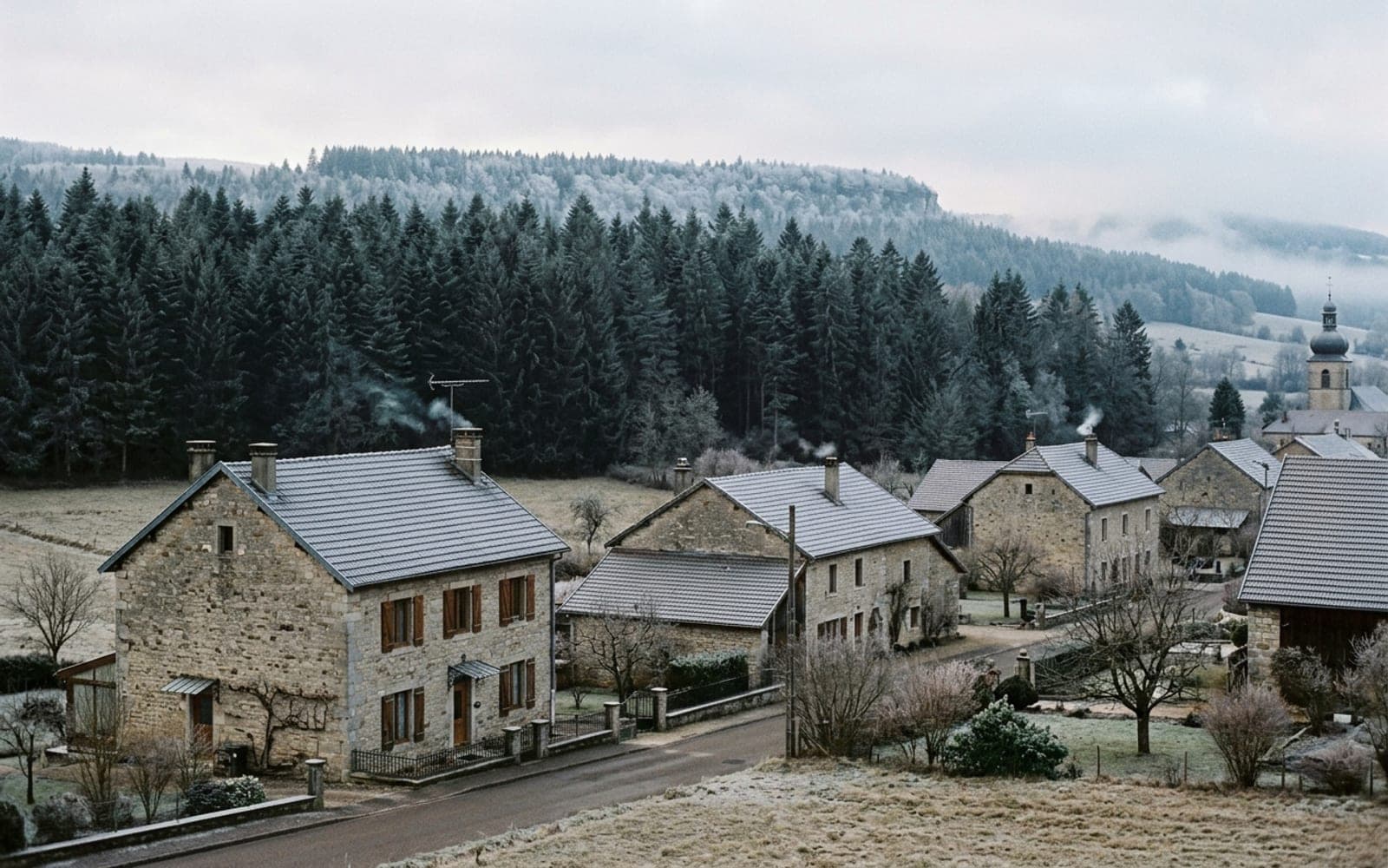Maisons de pierre en lisière d’une forêt de sapins givrée, fumée aux cheminées un matin d’hiver